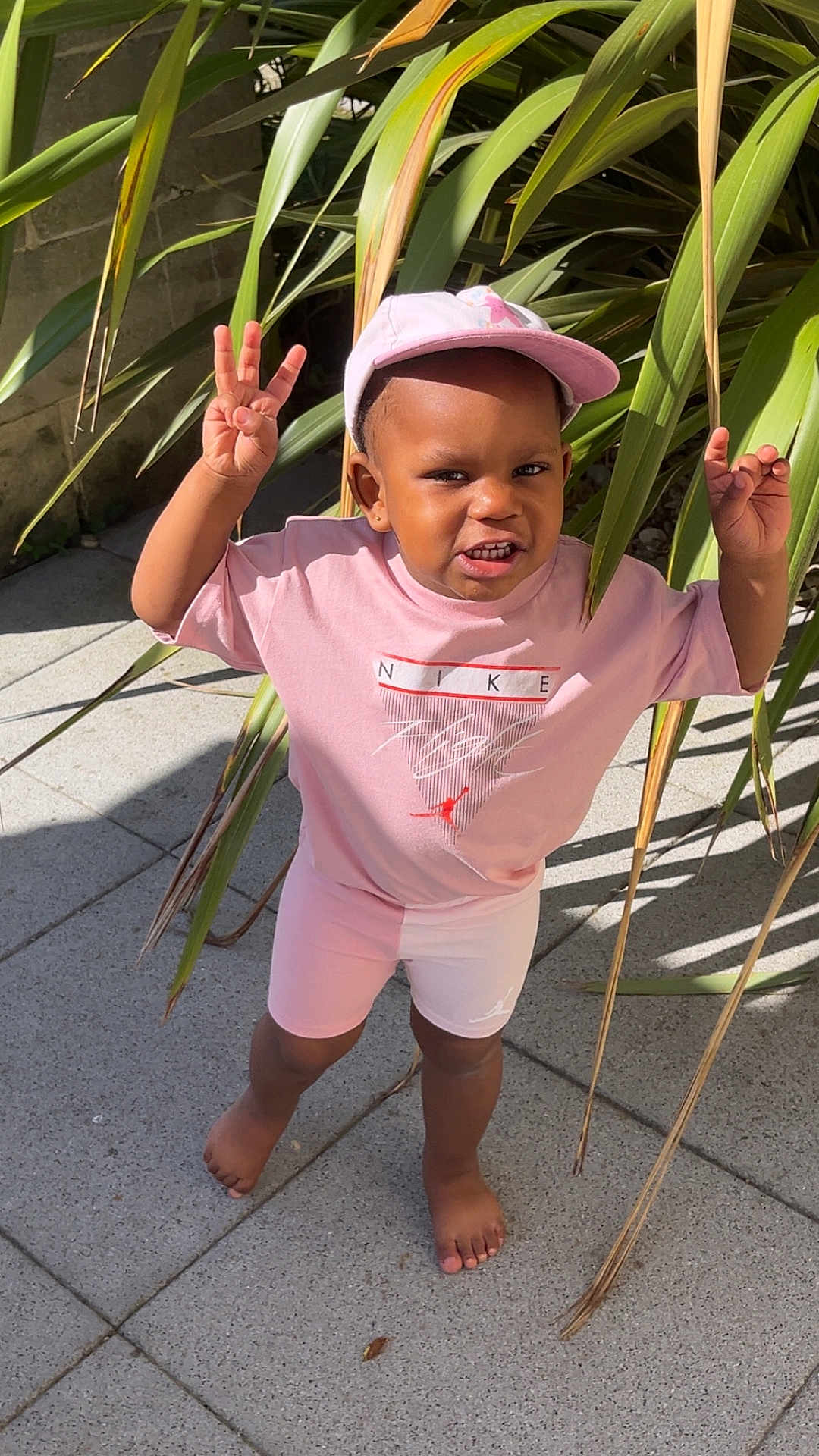 Loisse a rejoint le concours — aidez-le/la à gagner de superbes lots ! barefoot, cap, casual, child, concrete, gesture, greenery, happy, nike, outdoor, person, pink_clothing, plants, playful, shadow, sidewalk, smiling, sunlight, toddler, young_child