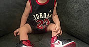 Mateo is registered to the contest to win money with this photo: baby, child, sitting, couch, sneakers, headband, jersey, clothing, person, indoor, footwear, cute, sportswear, portrait, young_child, fashion, red, black, white, casual
