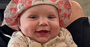 Leilani is registered to the contest to win money with this photo: baby, child, closeup, couch, cute, decor, face, floral_hat, furniture, hand, indoor, infant, joyful, living_room, onesie, person, plant, shelf, smiling, television