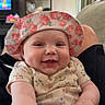 baby, child, closeup, couch, cute, decor, face, floral_hat, furniture, hand, indoor, infant, joyful, living_room, onesie, person, plant, shelf, smiling, television
