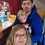 family, indoor, child, man, woman, glasses, blue_hoodie, table, plant, wooden_chair, curtain, potted_plant, casual_clothing, seated, portrait, people, young_child, home, natural_light, candid