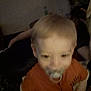 toddler, pacifier, child, orange_shirt, indoor, blurry, close_up, dim_light, person, young_child, infant, face, head, baby, portrait, young, human, indoors, people, clothing