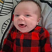 Liam is registered to the contest to win money with this photo: baby, infant, smile, tongue, red_clothing, black_clothing, checkered_pattern, blanket, leaf_pattern, happy, face, head, cute, young_child, lying_down, indoors, warm_clothing, closeup, person, child