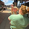 baby, bench, building, casual_clothing, child, cloud, daytime, glasses, hat, outdoor, person, reflection, shopping_center, sidewalk, sky, street, sunlight, waving, window, woman