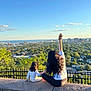 child, woman, cityscape, sky, trees, stone_ledge, bag, outdoor, nature, urban, horizon, sunlight, casual_clothing, back_view, fence, holding_hands, waving, greenery, blue_sky, people