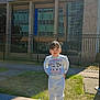 child, outdoor, building, stone_ledge, grass, sunlight, sweater, pants, sneakers, fence, window, concrete, person, smiling, daylight, hair_buns, casual_clothing, playful, young, urban