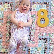 Johanna joined the competition — help win amazing prizes! baby, child, smiling, headband, romper, purple, floral_pattern, quilt, patchwork, colorful, number_eight, milestone, happy, infant, cute, lying_down, indoors, playful, celebration, person