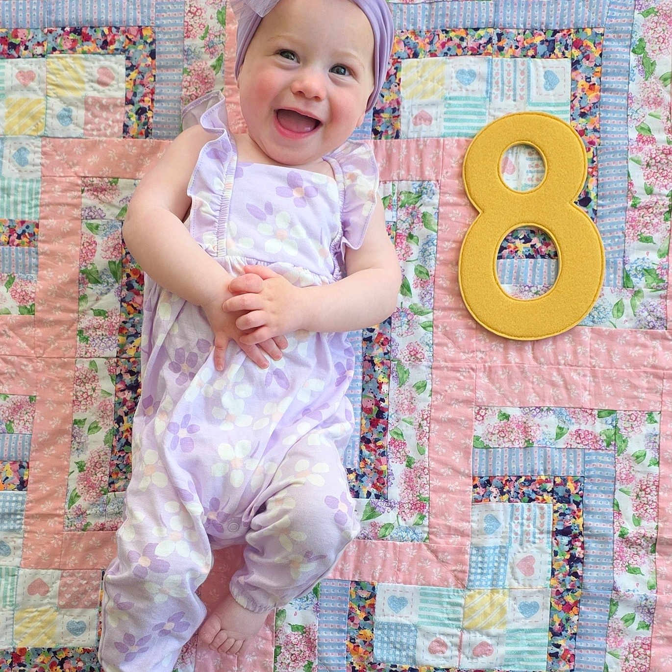 Johanna joined the competition — help win amazing prizes! baby, child, smiling, headband, romper, purple, floral_pattern, quilt, patchwork, colorful, number_eight, milestone, happy, infant, cute, lying_down, indoors, playful, celebration, person