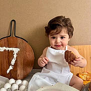 Jayce is registered to the contest to win money with this photo: toddler, child, apron, baking, flour, eggs, pastry, cookie, wooden_board, kitchen, cute, sitting, food, young_child, face, person, indoor, expression, holding, table
