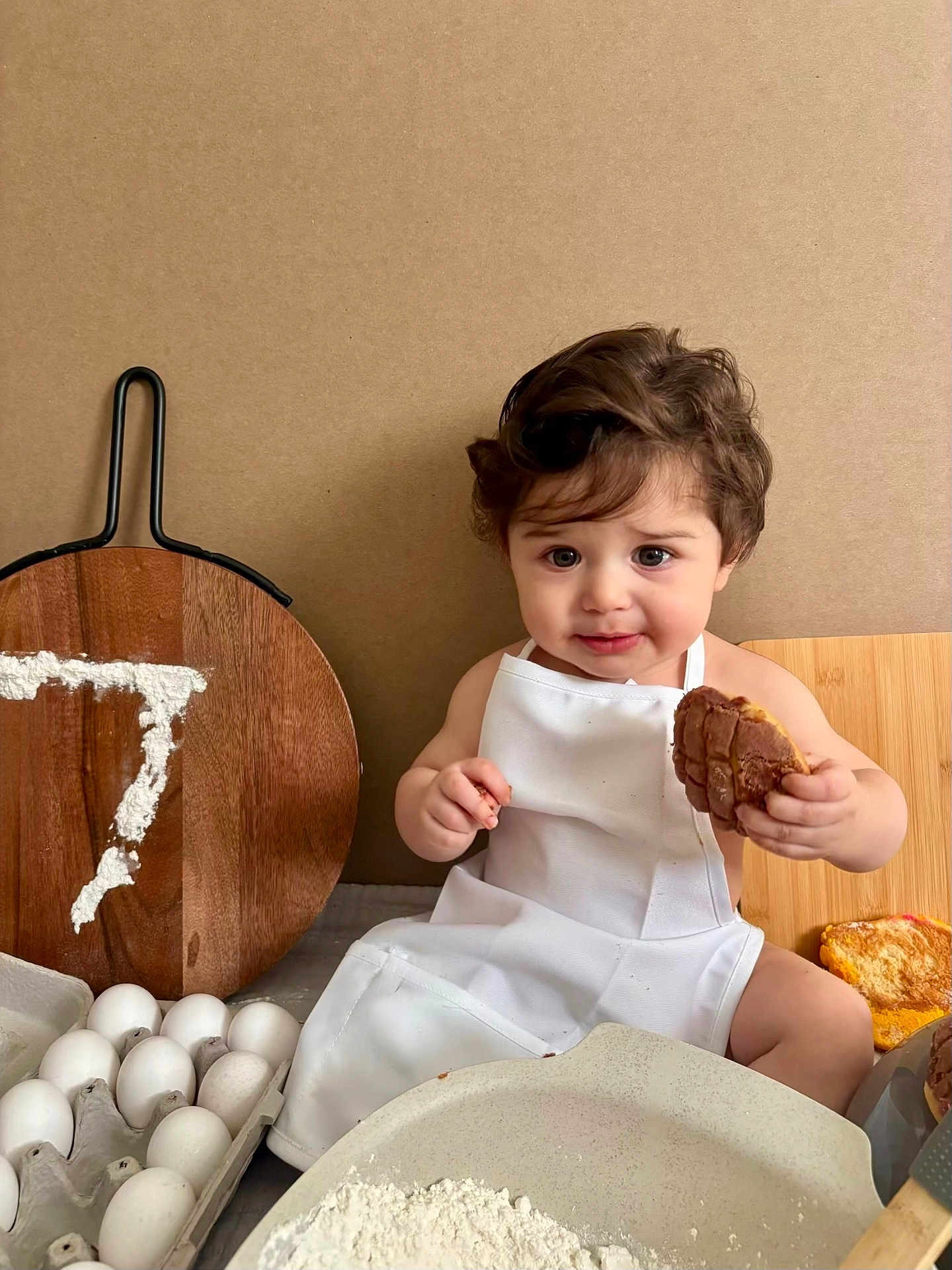 Jayce is registered to the contest to win money with this photo: toddler, child, apron, baking, flour, eggs, pastry, cookie, wooden_board, kitchen, cute, sitting, food, young_child, face, person, indoor, expression, holding, table