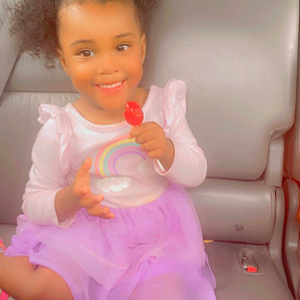 Ionia joined the competition — help win amazing prizes! car_seat, casual_clothing, child, cute, girl, hair, happy, indoor, joyful, lollipop, pigtails, portrait, purple_skirt, rainbow_shirt, seatbelt, sitting, smiling, sweet, toddler, young_child