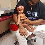 Reigh'Lynn joined the competition — help win amazing prizes! baby, man, child, smile, headband, tattoo, shirt, sitting, chair, floor, medical_room, bin, red_bin, cross_necklace, white_pants, colorful_sock, skin, indoor, person, holding