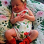 baby, sleeping, infant, onesie, headband, bow, red_booties, cushion, floral_pattern, peaceful, cute, hands, legs, indoors, soft_lighting, newborn, person, portrait, resting, cozy
