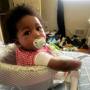 Amirah is registered to the contest to win money with this photo: baby, baby_seat, bedroom, blanket, brown_eyes, child, cozy, curly_hair, cushion, indoors, infant, looking_at_camera, natural_light, pacifier, plaid_clothing, portrait, sitting, smiling, toy, window