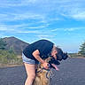 person, dog, outdoor, sky, hill, path, greenery, affection, hug, pet, animal, casual_clothing, shorts, tshirt, leash, nature, summer, smiling, happy, friendship