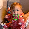 Azmae is registered to the contest to win money with this photo: baby, child, infant, face, portrait, sitting, playpen, teething_ring, toy, pajamas, pink_clothing, red_hair, wide_eyes, indoors, cushion, blanket, colorful, baby_gear, play_area, smiling