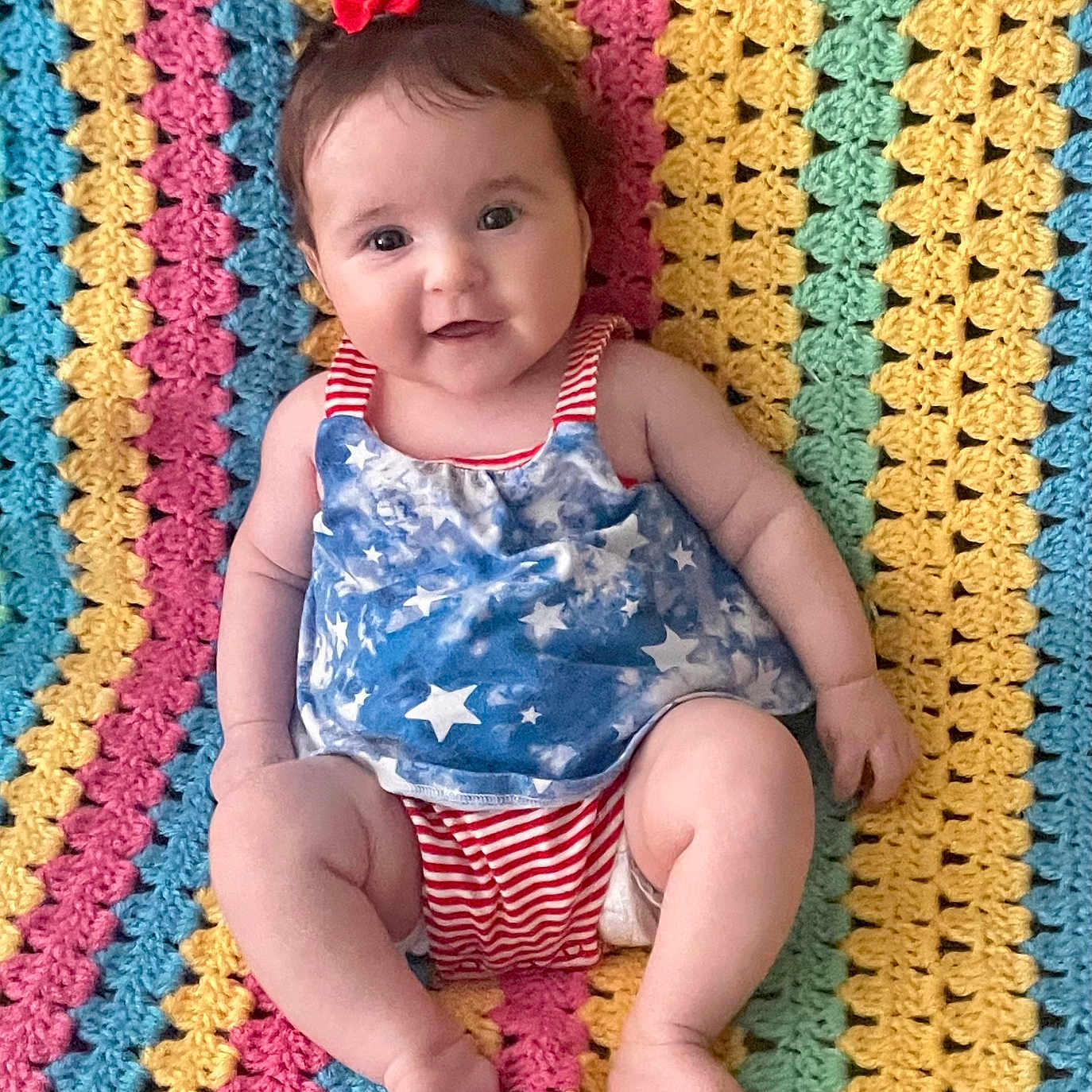 Rosie is registered to the contest to win money with this photo: baby, child, infant, cute, smiling, colorful_blanket, crocheted_blanket, red_bow, blue_top, striped_clothing, laying_down, happy, portrait, indoors, soft_texture, patterned, small_person, skin, legs, arms