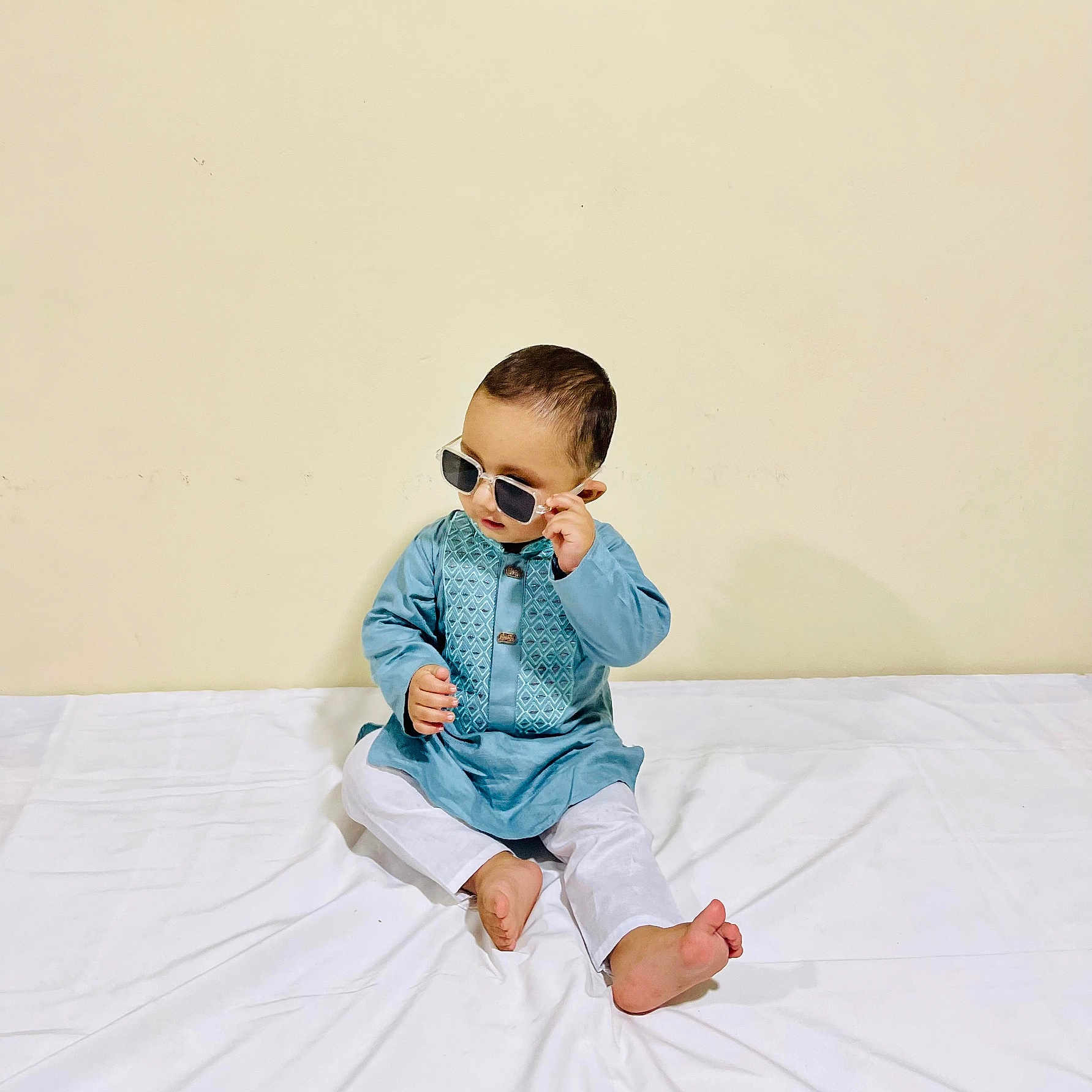 Mihad Rahman is registered to the contest to win money with this photo: baby, barefoot, blue_clothing, child, curious, cute, face, fashion, head, indoor, minimalist_background, person, plain_wall, portrait, sitting, small, sunglasses, traditional_clothing, white_pants, young_child