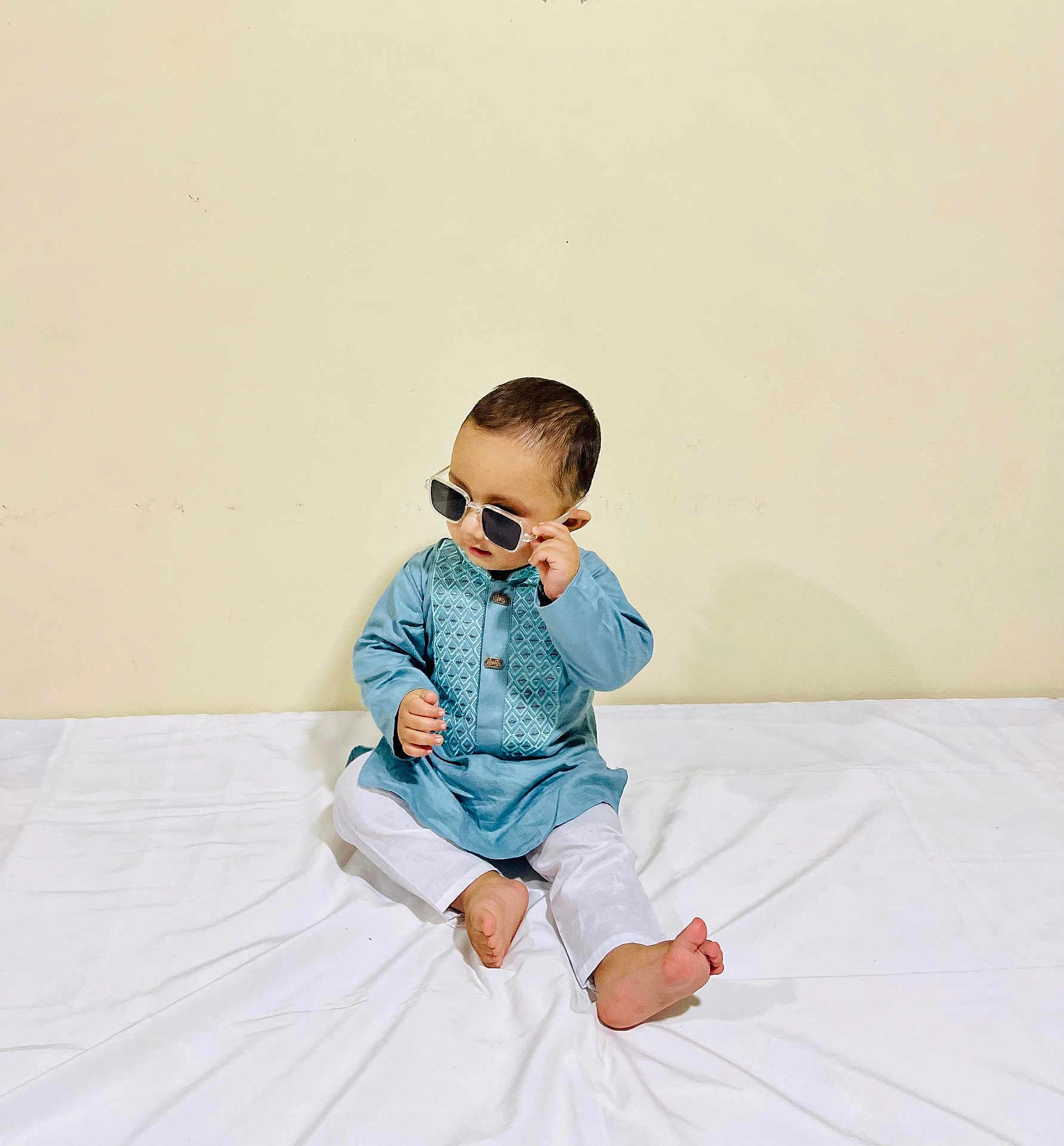 Mihad Rahman is registered to the contest to win money with this photo: baby, child, sunglasses, blue_clothing, white_pants, barefoot, sitting, curious, indoor, plain_wall, traditional_clothing, portrait, cute, young_child, fashion, minimalist_background, person, head, face, small