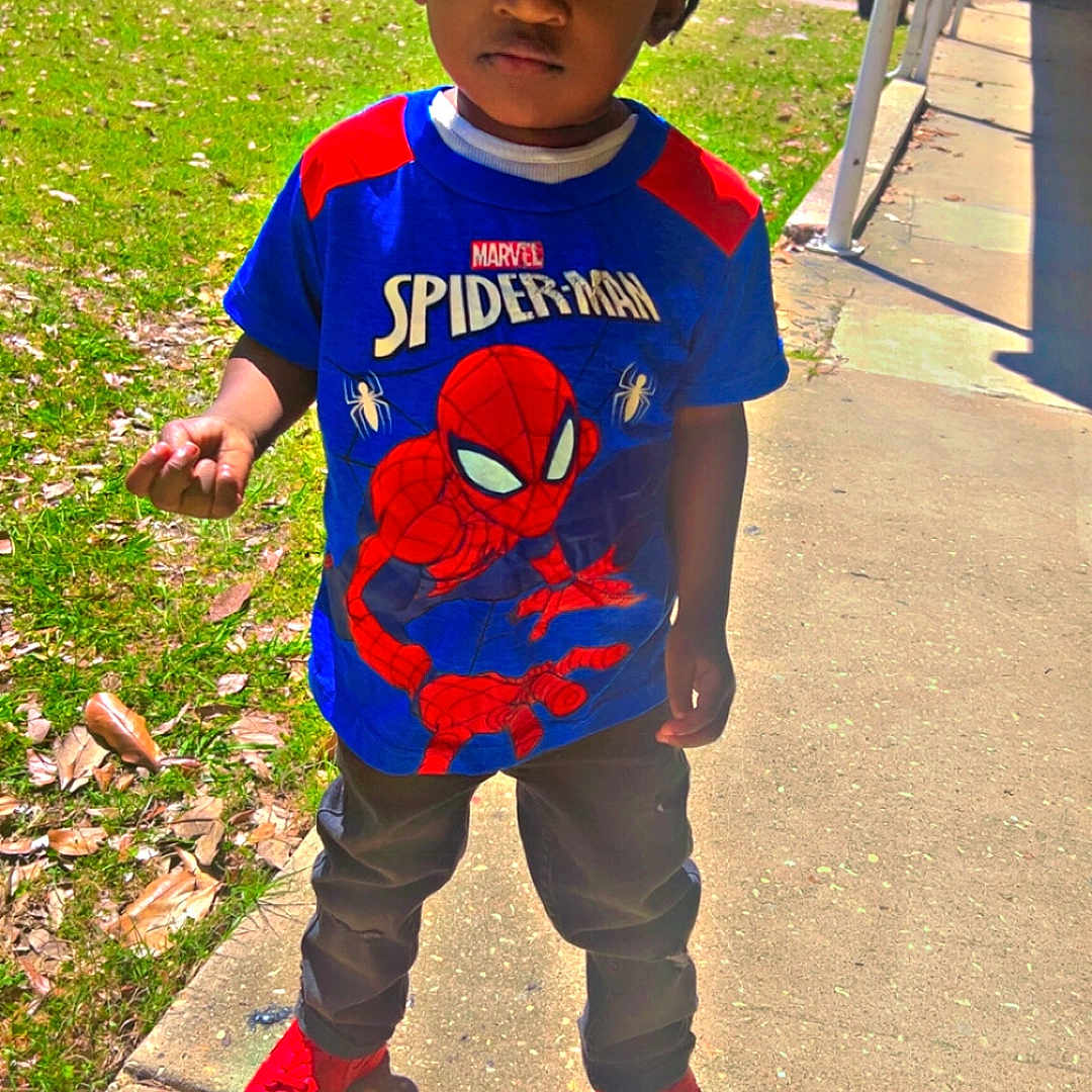 Zaiden is registered to the contest to win money with this photo: child, boy, outdoor, sidewalk, grass, tree, shirt, spiderman, red_shoes, shadow, daylight, casual_clothing, hair, expression, park, sunny, playground, young, person, standing