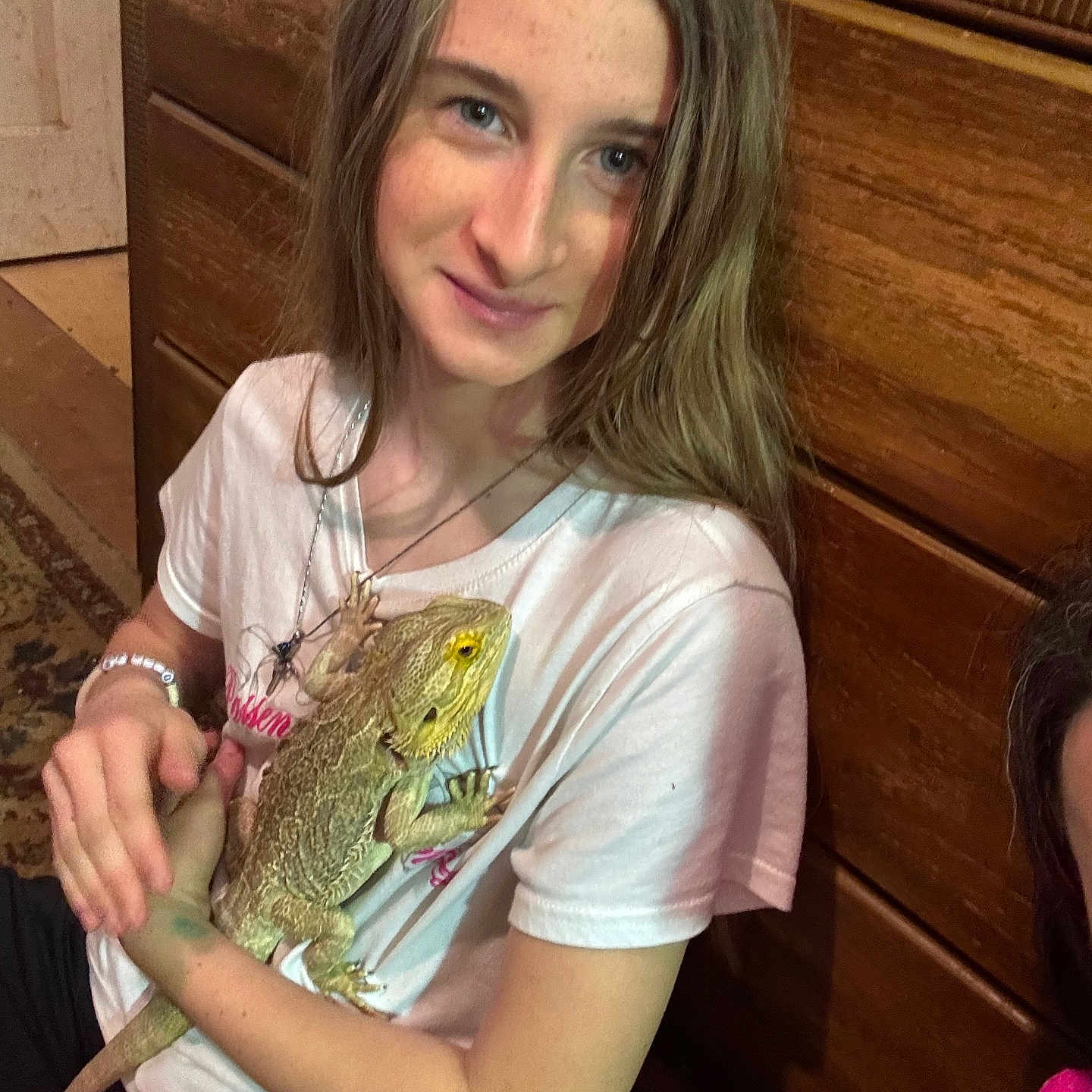 Amberlyn is registered to the contest to win money with this photo: child, girl, lizard, bearded_dragon, pet, white_tshirt, bracelet, necklace, long_hair, smile, indoor, wooden_furniture, reptile, animal, person, casual_clothing, young, sitting, holding, friendly