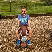 Hunter is registered to the contest to win money with this photo: child, boy, playground, spring_rocker, wood_chips, grass, outdoor, smiling, blue_shoes, casual_clothing, short_hair, daylight, fence, recreation, fun, happy, summer, toy, park, nature