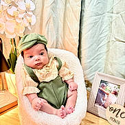 Aiden is registered to the contest to win money with this photo: baby, infant, child, green_clothing, hat, chair, flower, orchid, photo_frame, milestone, one_month, wooden_surface, curtains, indoor, portrait, cute, cozy, soft_lighting, smiling, sitting