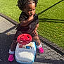 child, toddler, afro_hairstyle, pink_sneakers, toy_car, police_car_toy, outdoor, playground, grass, pavement, sunlight, shadow, black_dress, curious, sitting, finger_in_mouth, young_child, playtime, daytime, casual_clothing