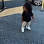 toddler, child, standing, asphalt, parking_lot, car, black_shirt, beige_pants, white_shoes, curly_hair, urban, outdoor, daylight, shadow, fence, vehicle, side_view, casual_clothing, person, young_child