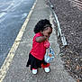 toddler, child, sidewalk, red_shirt, black_skirt, white_shoes, purse, toy, curly_hair, outdoor, urban, brick_wall, mulch, street, casual_clothing, young_child, standing, playful, earrings, daylight
