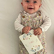 Ninon participe au concours pour gagner de l'argent avec cette photo : baby, infant, smiling, floral_romper, card, hand, face, blue_eyes, lying_down, white_background, cute, child, person, happy, clothing, button, skin, head, portrait, baby_girl