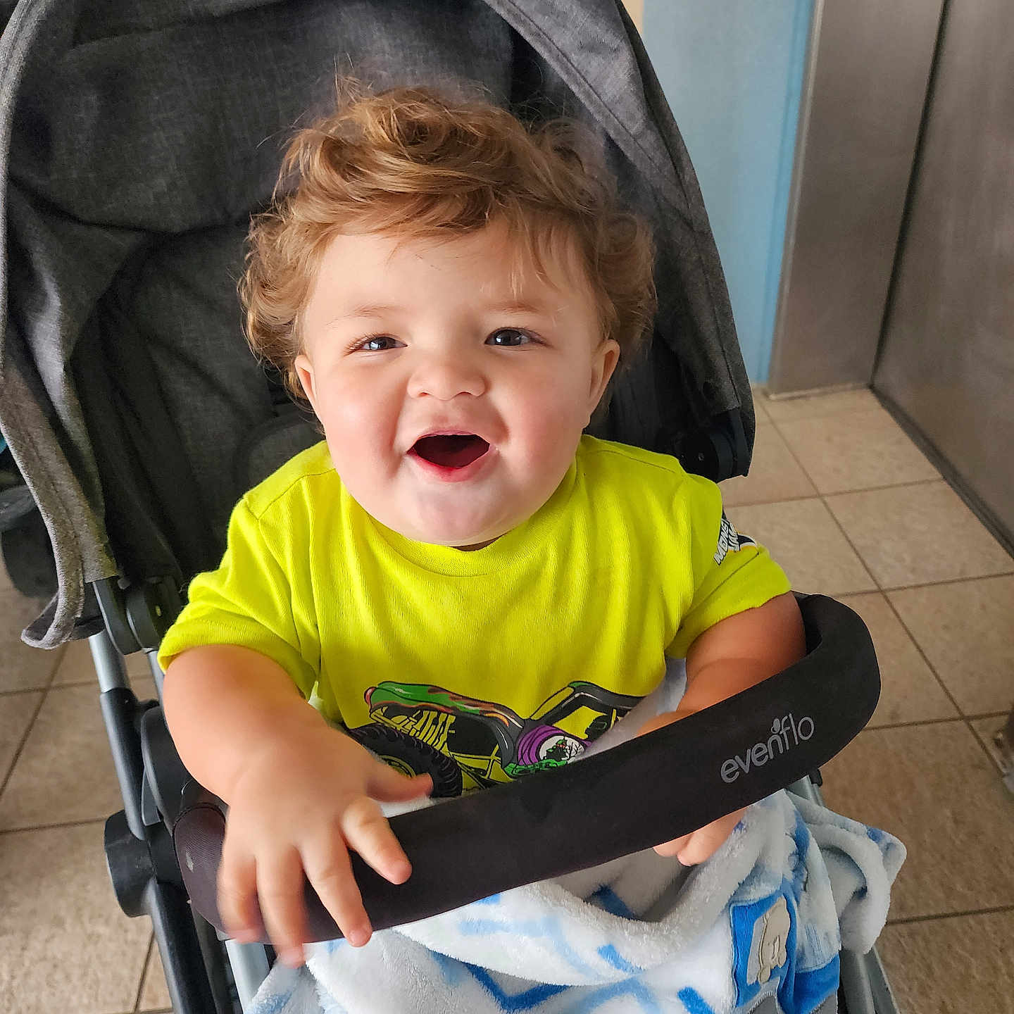 Xander is registered to the contest to win money with this photo: toddler, child, stroller, yellow_shirt, blanket, indoor, happy, curly_hair, smiling, baby, person, floor_tiles, elevator, hand, clothing, seat, toy_car_print, bright_colors, facial_expression, grip