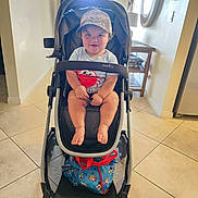 Xander joined the competition — help win amazing prizes! toddler, stroller, baby, child, cap, indoor, tile_floor, person, smiling, seated, clothing, table, bag, adult, background, hat, footwear, furniture, light, family