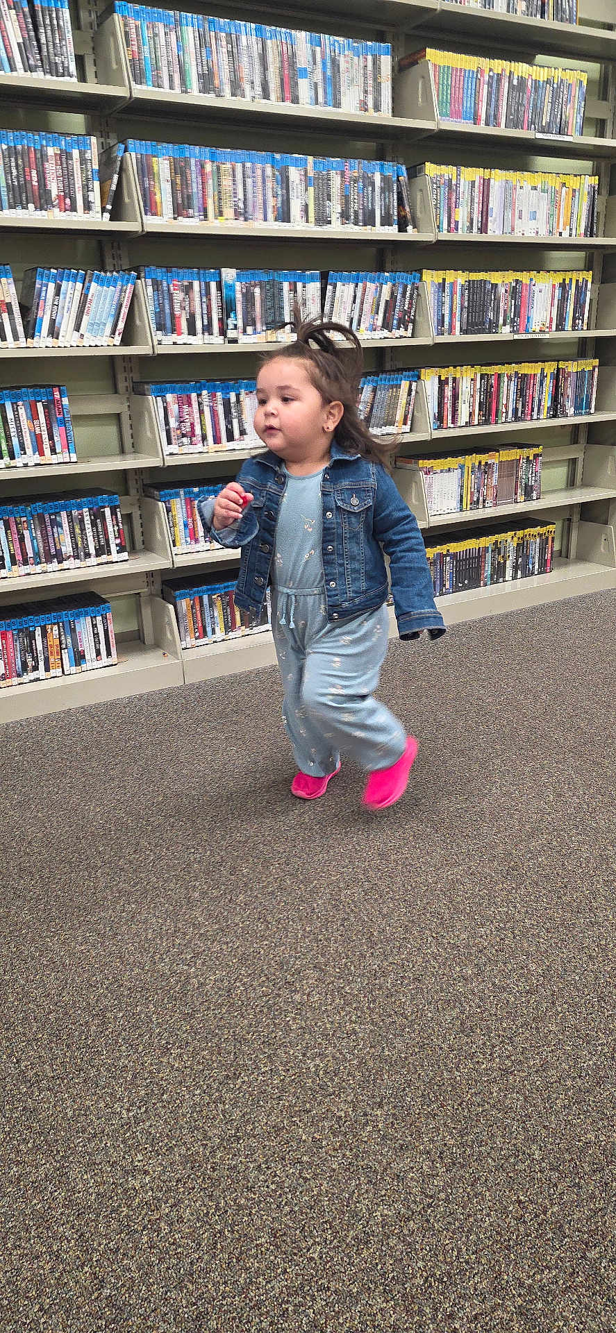 Ruby is registered to the contest to win money with this photo: toddler, child, running, denim_jacket, blue_jumpsuit, pink_shoes, carpet, bookshelf, library, indoor, person, young_child, hair, clothing, shelf, floor, motion, aisle, casual, cute