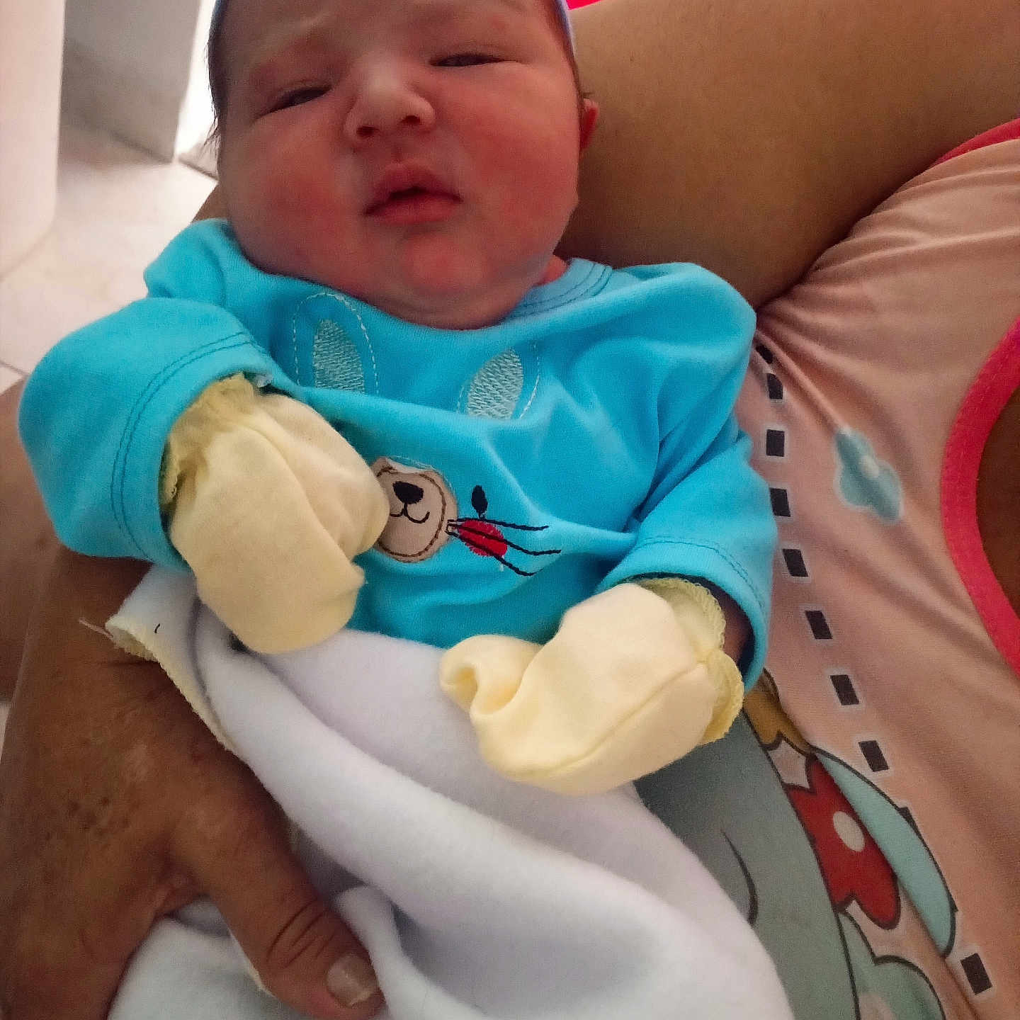 Thiago Perez is registered to the contest to win money with this photo: adult_hand, baby, blanket, blue_clothing, cozy, cute, face, headwear, holding, indoor, infant, mittens, newborn, person, pink_background, portrait, sleepy, soft, tender_moment, young_child