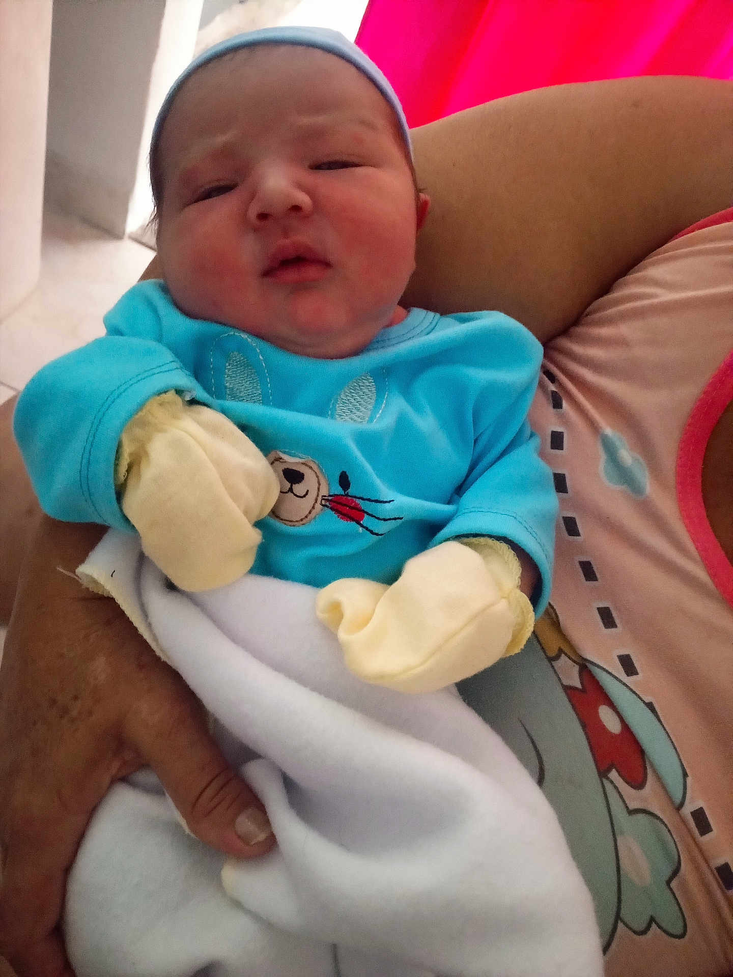 Thiago Perez is registered to the contest to win money with this photo: baby, newborn, infant, mittens, blue_clothing, blanket, adult_hand, face, person, indoor, holding, pink_background, portrait, cute, sleepy, young_child, headwear, cozy, soft, tender_moment