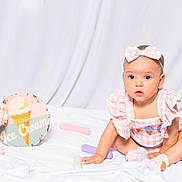 Davae is registered to the contest to win money with this photo: baby, child, clothing, curious, cute, decor, gingham, headband, ice_cream_sign, indoor, infant, looking, pastel_colors, pink_bow, playtime, portrait, sitting, soft_lighting, toys, white_sheet