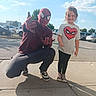Raelynn is registered to the contest to win money with this photo: child, spiderman, superhero, costume, mask, smiling, thumbs_up, outdoor, parking_lot, cars, sidewalk, sunny, sky, clouds, portrait, standing, crouching, tshirt, sneakers, playful