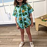 blue_wall, brown_hair, casual_clothing, child, fashion, floral_shirt, full_body, happy, home, indoor, playroom, portrait, smiling, sneakers, standing, text_overlay, toy_table, tropical_pattern, white_paneling, wooden_floor