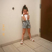 Raelynn is registered to the contest to win money with this photo: child, girl, toddler, standing, portrait, indoor, hallway, door, tile_floor, white_shirt, striped_shorts, sandals, hair_bun, finger_guns, playful, pose, smiling, timestamp, wall, full_body