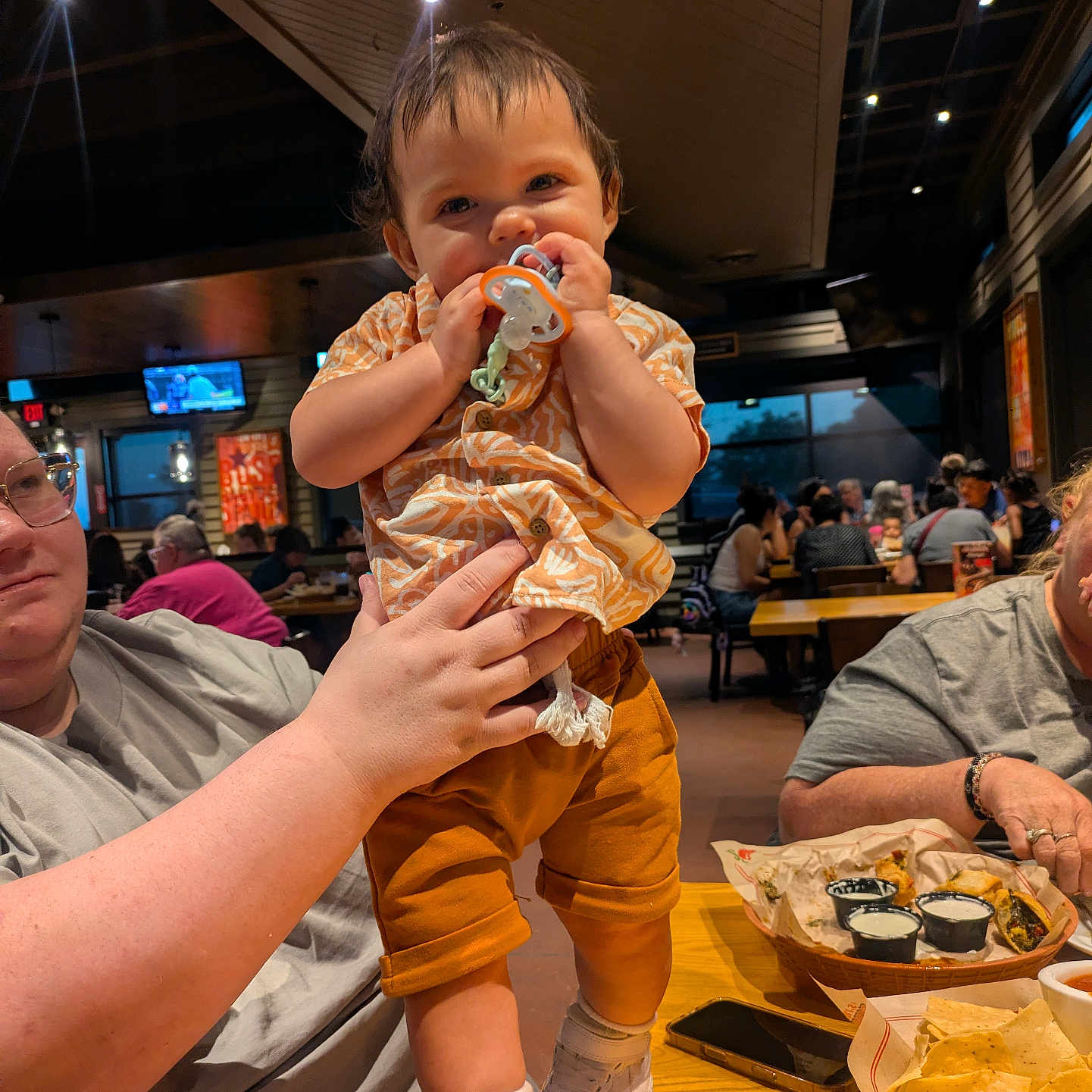 Toebias is registered to the contest to win money with this photo: baby, child, infant, restaurant, dining, pacifier, smiling, standing, adult, family, table, food, tacos, tortilla_chips, dipping_sauce, smartphone, indoor, ceiling_lights, casual_dining, portrait