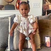 Chloe is registered to the contest to win money with this photo: baby, child, clothing, couch, cushion, decor, expression, face, footwearless, hair_bows, indoor, living_room, pacifier_clip, patterned_outfit, person, phone_screen, striped_cushion, toddler, video_call, wall_switch