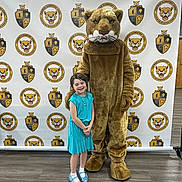 Lilly joined the competition — help win amazing prizes! child, girl, mascot, cougar, school, elementary_school, background, floor, blue_dress, smiling, costume, indoor, footwear, person, posing, event, logo, photography, happy, standing