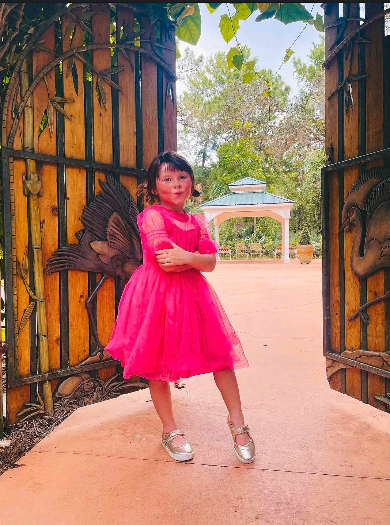 Lilly is registered to the contest to win money with this photo: child, girl, pink_dress, pose, wooden_gate, carving, bird_design, outdoor, gazebo, greenery, trees, playful, smile, shoes, pigtails, daylight, pathway, nature, cute, fashion