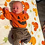 baby, blanket, brown_pants, child, cute, decor, fall_theme, floor, happy, holiday, indoor, infant, laying_down, newborn, orange_clothing, pattern, person, pumpkin, socks, soft_texture