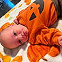 autumn_leaves, baby, blanket, child, cozy, curious_expression, cute, face, fist, head, indoors, infant, lying_down, orange_clothing, portrait, pumpkin_design, skin, soft_texture, warm_colors, young