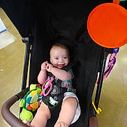 Sylvia is registered to the contest to win money with this photo: baby, stroller, toy, smile, child, infant, happy, seatbelt, black, colorful, play, indoor, person, cute, hand, foot, clothing, green, pink, orange
