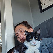 Pepper is registered to the contest to win money with this photo: person, cat, indoor, smiling, black_and_white_cat, portrait, sketch, wall_art, window, natural_light, holding, relaxed, furniture, gray_shirt, hair_up, whiskers, face, ears, couch, hand