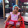 baby, blue_headband, bracelet, child, clothing, curious, expression, floor, footwear, furniture, headwear, indoor, mesh_screen, person, pink_dress, playground, portrait, red_chair, sitting, toddler