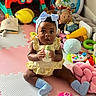 baby, blue_bow, bottle, child, colorful, cute, expression, floor, fun, headband, indoor, infant, playmat, playtime, plush_toys, room, sitting, socks, toys, yellow_dress